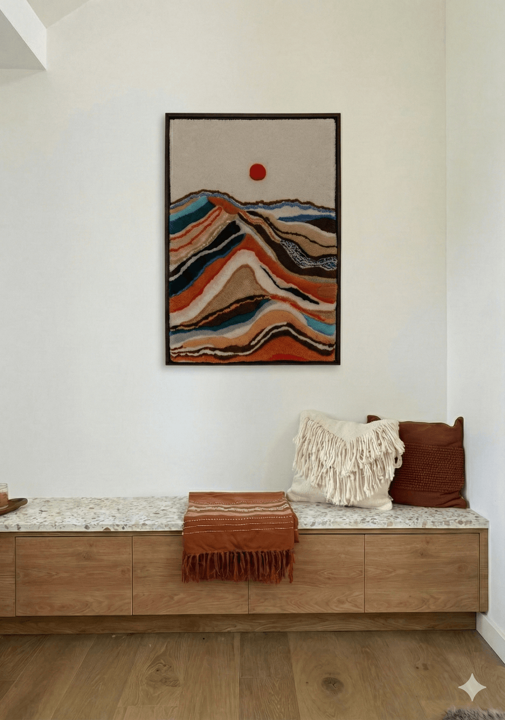 Handmade tufted wall hanging with colorful layered mountain landscape and red sun, displayed above a modern oak bench with terrazzo top
