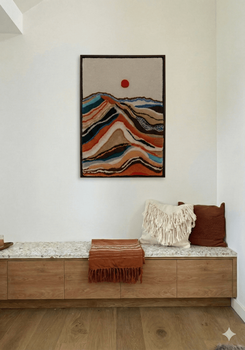 Handmade tufted wall hanging with colorful layered mountain landscape and red sun, displayed above a modern oak bench with terrazzo top