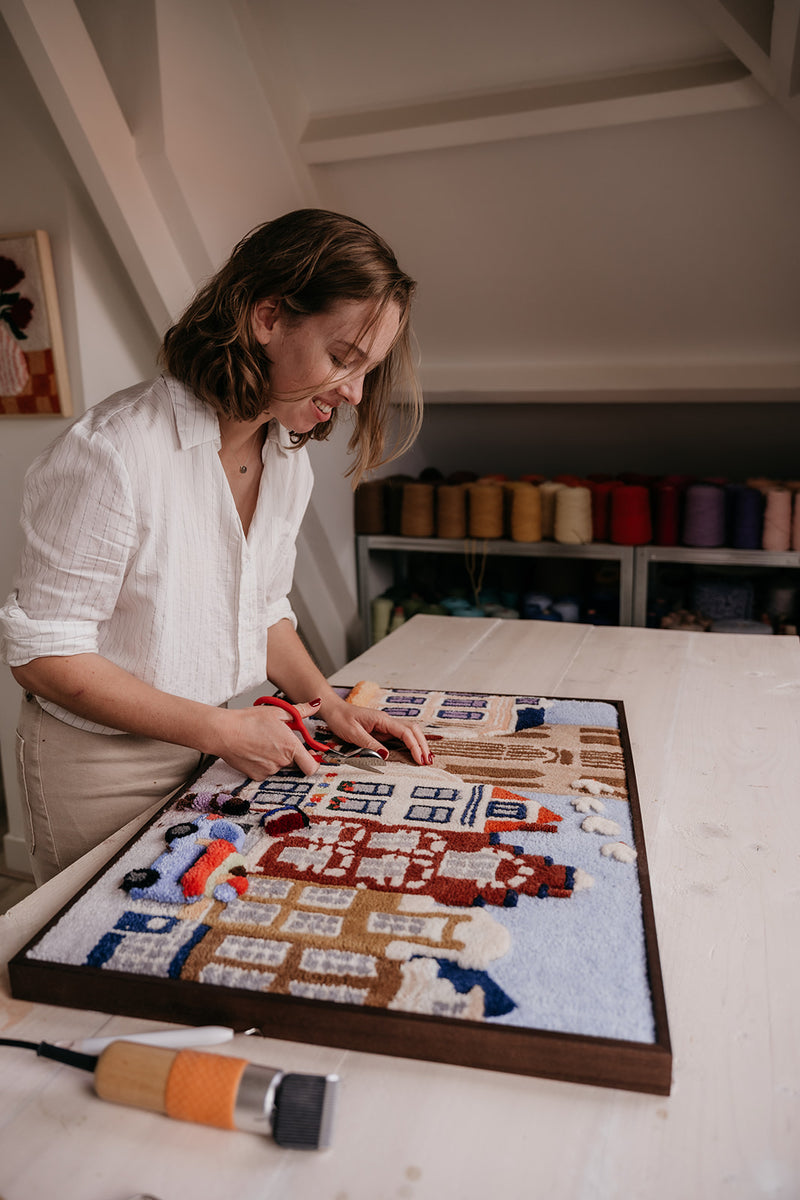 Textile artist Martine Schulten trimming a custom tufted wall hanging of colorful Dutch canal houses with scissors at her worktable in the Filo Arts studio in Utrecht