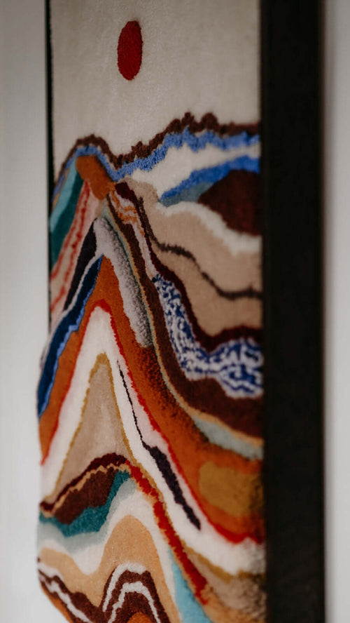 Close-up side view of tufted wall hanging Dawn showing the layered wool texture of colorful mountain ridges with red sun in a dark wooden frame