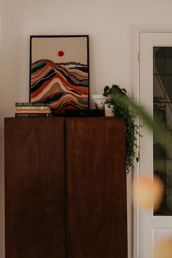 Tufted wall hanging Dawn by Filo Arts with colorful mountain layers and red sun, styled on a vintage walnut cabinet with books and trailing plant
