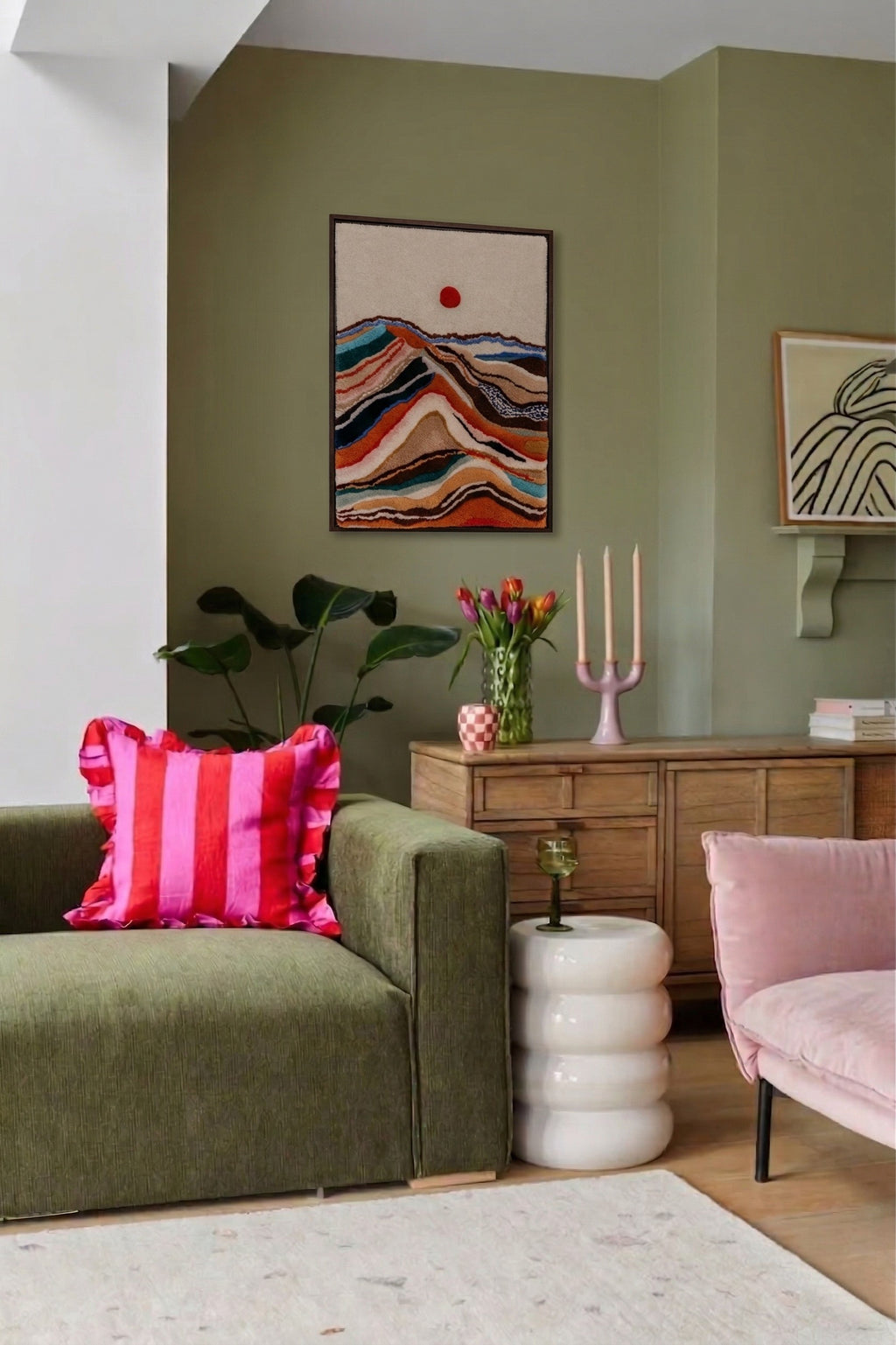 Hand-tufted wall hanging depicting a layered mountain landscape with a red sun, displayed in a sage green living room above a wooden sideboard
