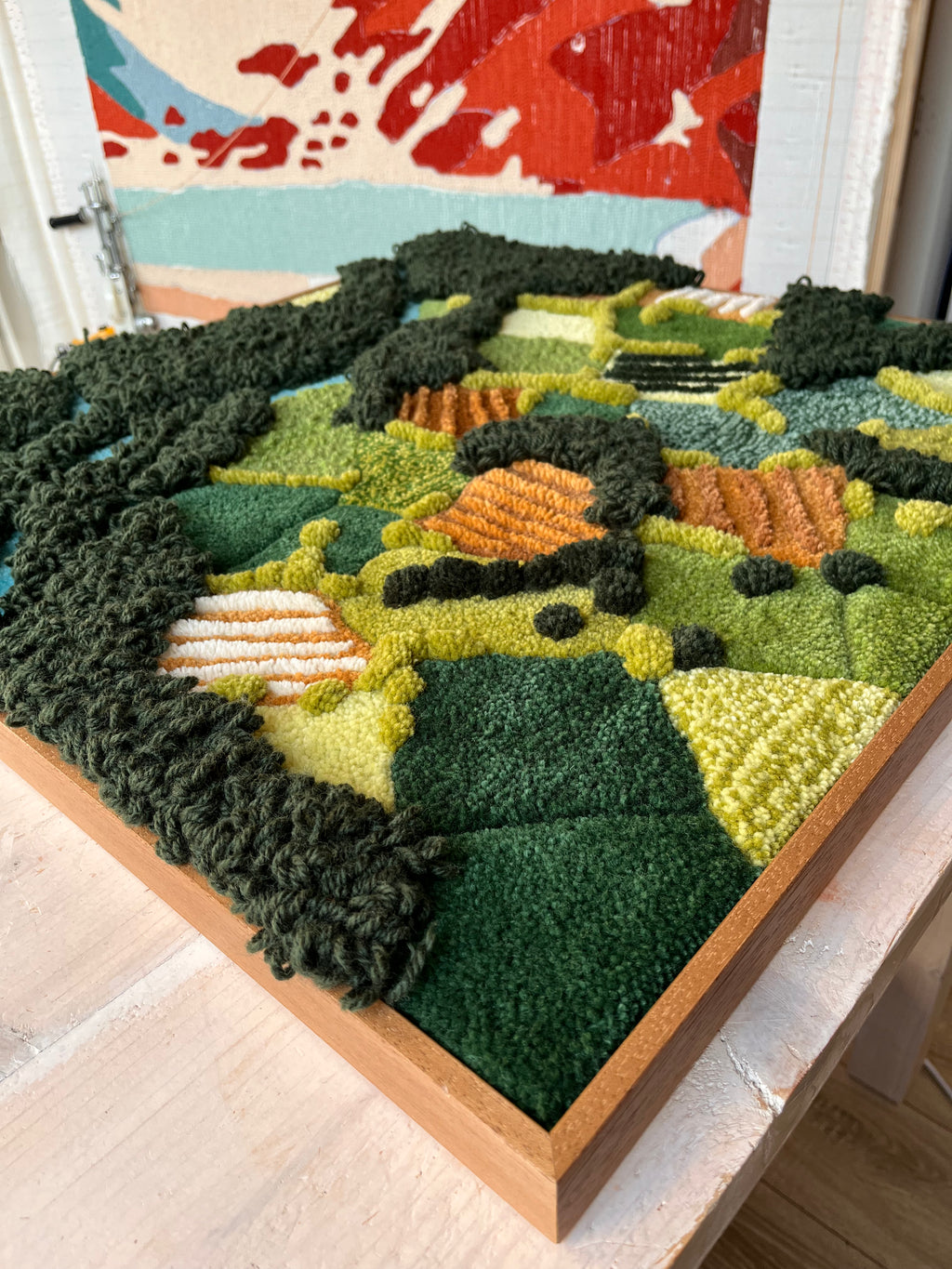 Close-up of handmade tufted wall hanging Maedow showing textured wool landscape in shades of green, yellow and orange in a wooden oak frame