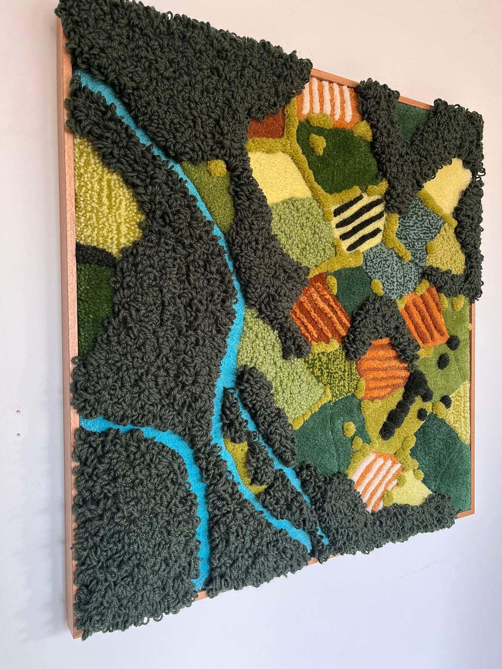 Side view of tufted wall hanging Meadow showing the 3D wool texture with raised dark green loops, blue river detail and orange accents in an oak frame
