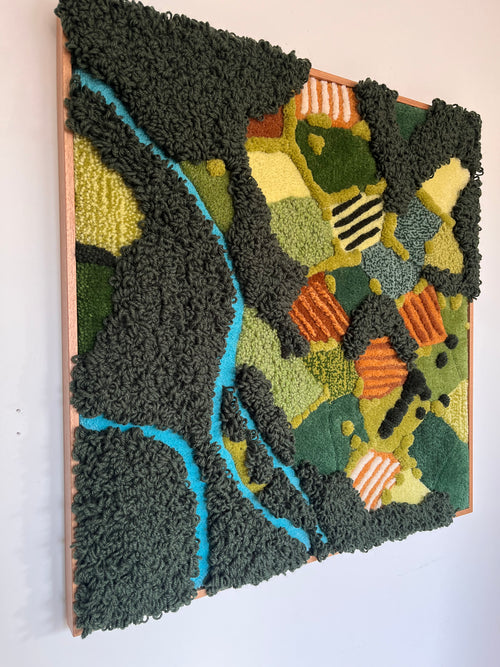 Side view of tufted wall hanging Meadow showing the 3D wool texture with raised dark green loops, blue river detail and orange accents in an oak frame