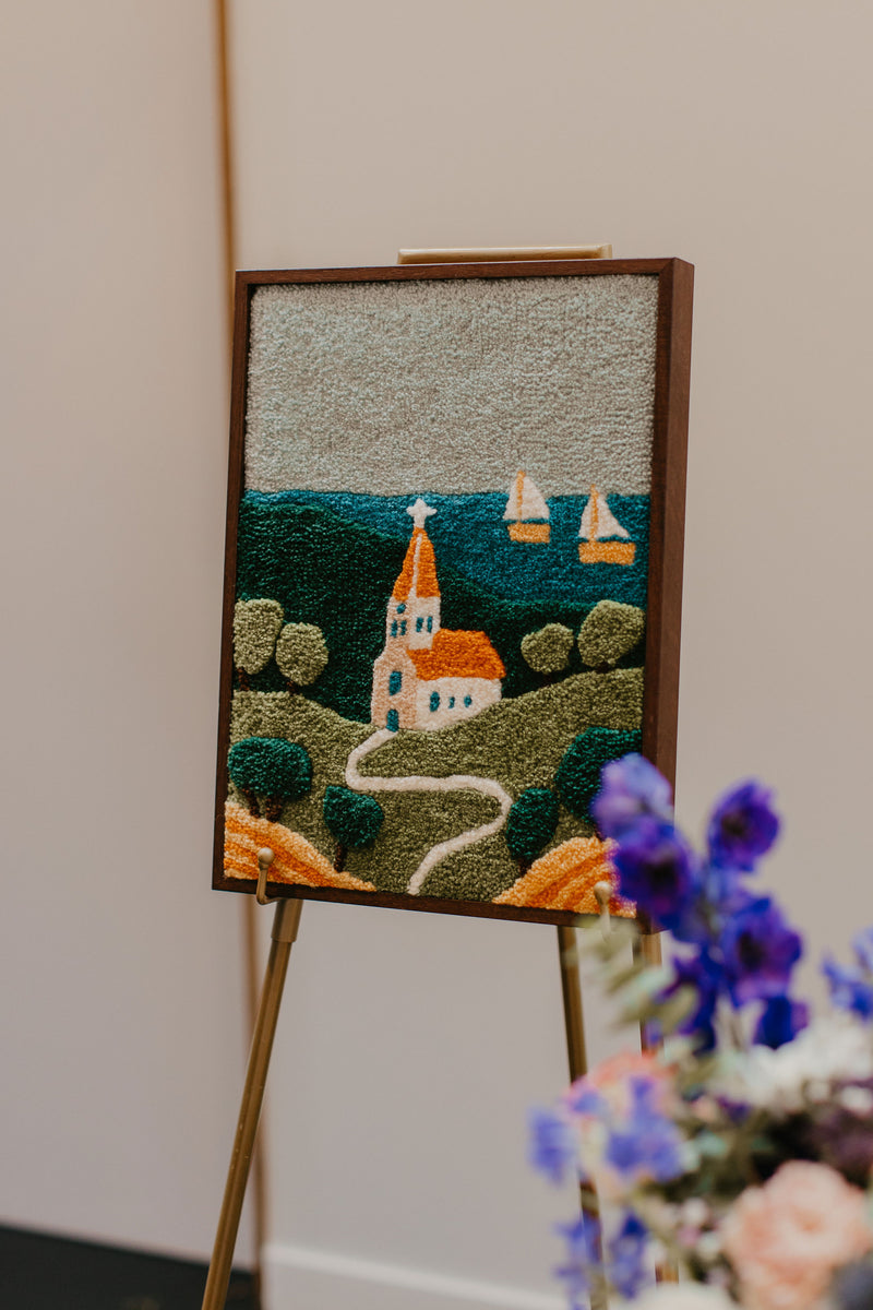 Custom tufted wall hanging of a coastal village with church, winding path, green hills and sailboats on the sea, displayed on a gold easel at an event