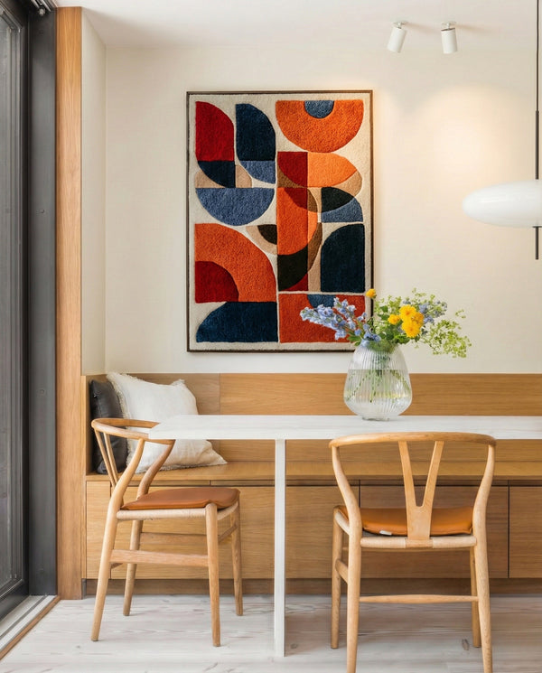 Hand-tufted wall hanging with bold geometric shapes in orange, red and navy blue, displayed above a built-in oak bench in a minimalist dining nook